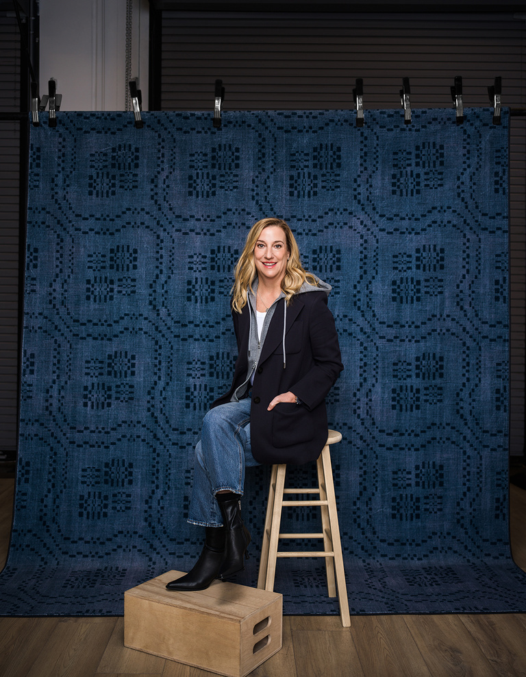 Nicole Otto sitting on a stool with a blue rug hung behind her as a backdrop