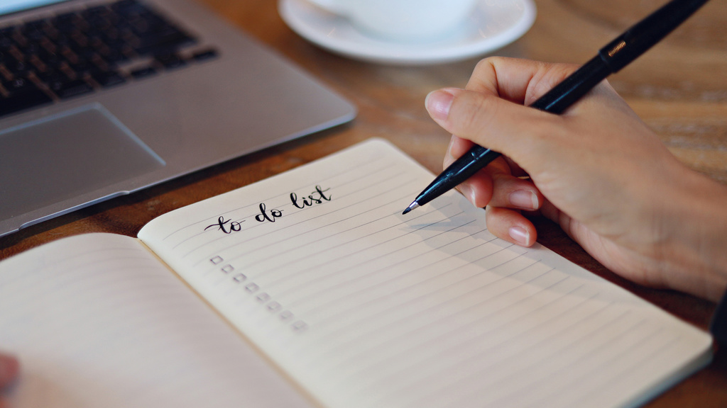 A pen in hand, writing a to do list on a pad of paper in front of a computer. 